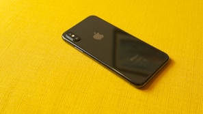 A black smartphone with a reflective surface lies on a vibrant yellow textured fabric. The device's back features a dual camera setup and a notable logo, with visible text at the bottom indicating its model.