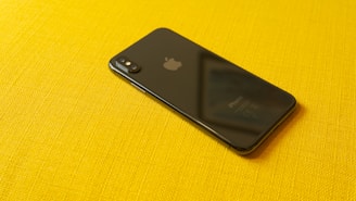A black smartphone with a reflective surface lies on a vibrant yellow textured fabric. The device's back features a dual camera setup and a notable logo, with visible text at the bottom indicating its model.