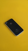 A black smartphone is resting on a bright yellow surface. The phone's back features a prominent silver apple logo, and its design is sleek and minimalist.