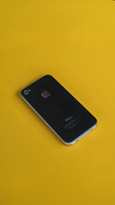 A black smartphone is resting on a bright yellow surface. The phone's back features a prominent silver apple logo, and its design is sleek and minimalist.
