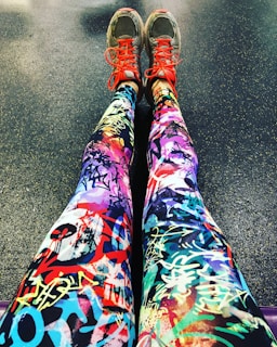 A pair of legs wearing colorful leggings with vibrant graffiti-like patterns, accompanied by gray sneakers that feature bright red laces. The background is a textured floor, possibly indicating a gym or workout area.