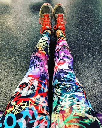 A pair of legs wearing colorful leggings with vibrant graffiti-like patterns, accompanied by gray sneakers that feature bright red laces. The background is a textured floor, possibly indicating a gym or workout area.