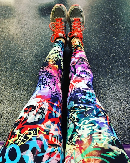 A pair of legs wearing colorful leggings with vibrant graffiti-like patterns, accompanied by gray sneakers that feature bright red laces. The background is a textured floor, possibly indicating a gym or workout area.