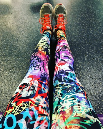 A pair of legs wearing colorful leggings with vibrant graffiti-like patterns, accompanied by gray sneakers that feature bright red laces. The background is a textured floor, possibly indicating a gym or workout area.