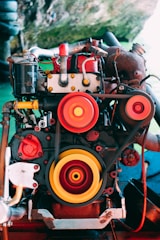 An industrial engine featuring prominent red and yellow pulleys is set against an outdoor environment. The intricate mechanical components are highlighted through vibrant colors, blending with the natural green backdrop.