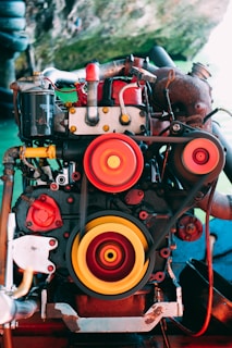 An industrial engine featuring prominent red and yellow pulleys is set against an outdoor environment. The intricate mechanical components are highlighted through vibrant colors, blending with the natural green backdrop.
