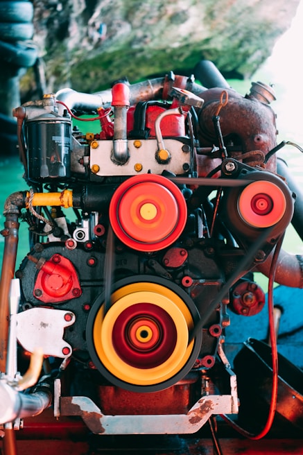 An industrial engine featuring prominent red and yellow pulleys is set against an outdoor environment. The intricate mechanical components are highlighted through vibrant colors, blending with the natural green backdrop.