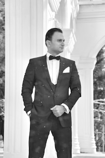 A man in a formal black suit and bow tie stands confidently with hands in pockets. The backdrop features classic architectural columns and arches, suggesting an elegant setting.