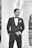 A man in a formal black suit and bow tie stands confidently with hands in pockets. The backdrop features classic architectural columns and arches, suggesting an elegant setting.
