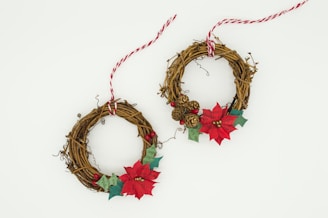 Two decorative wreaths made of intertwined brown twigs. Each wreath is embellished with red poinsettias, small ornamental berries, green leaves, and miniature pine cones. Red and white striped twine is tied around each wreath for hanging.