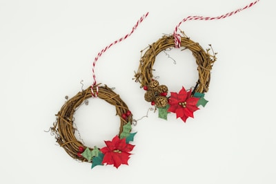 Two decorative wreaths made of intertwined brown twigs. Each wreath is embellished with red poinsettias, small ornamental berries, green leaves, and miniature pine cones. Red and white striped twine is tied around each wreath for hanging.