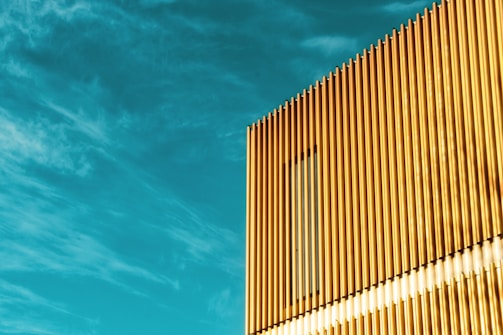 A modern building facade featuring vertical golden panels in strong sunlight, set against a vivid blue sky with sparse clouds.