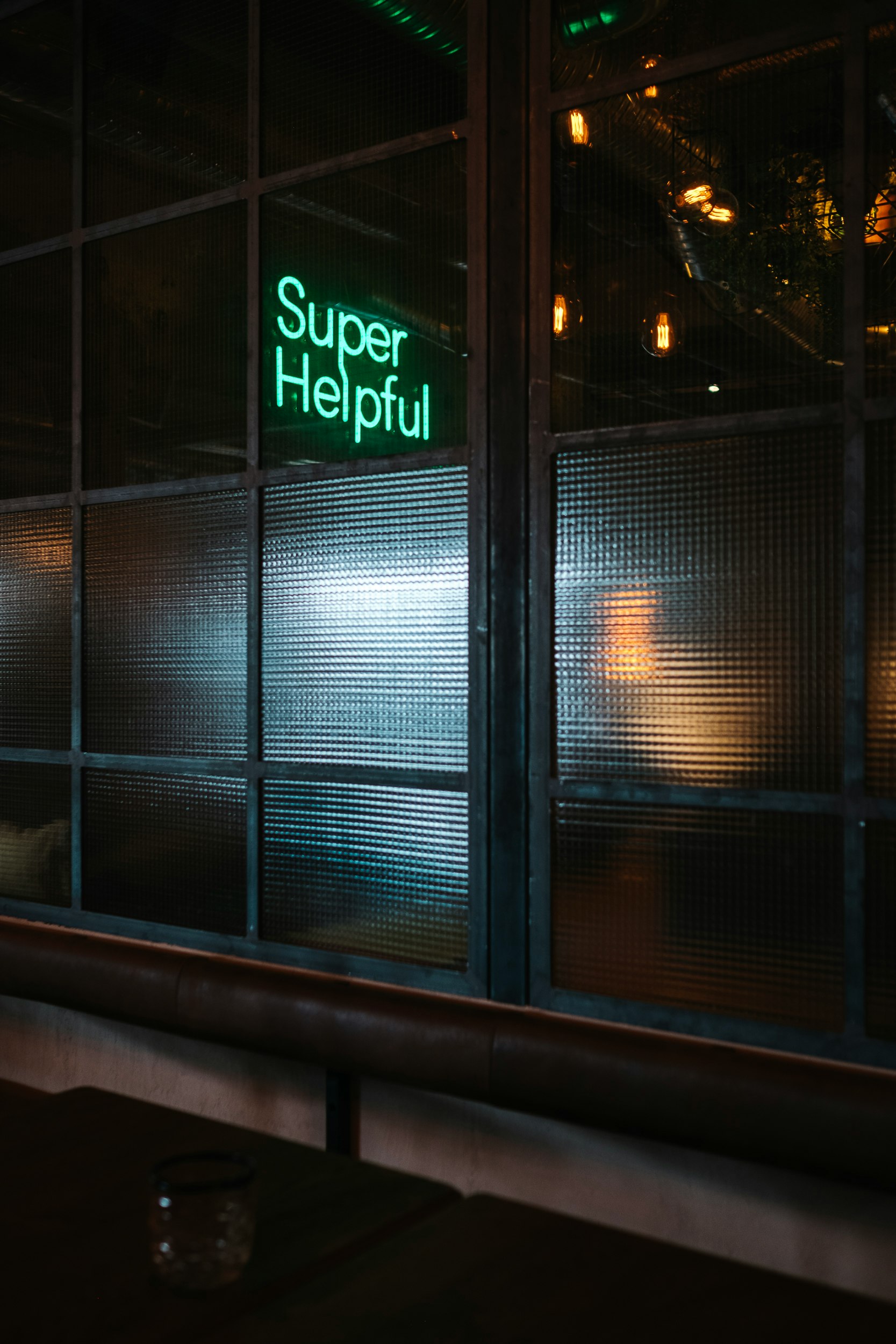 Photograph from the inside of a dimly lit Cafe, at the window, which contains a neon sign with the text "Super Helpful."