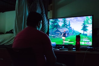 A person is sitting in front of a computer monitor, playing a video game. The room is dimly lit, likely with natural light seeping in slightly, and a bed is visible in the background. The game on the screen shows a character running through a green, forested area.