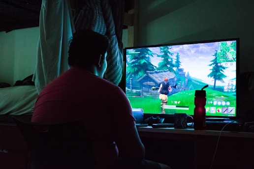 A person is sitting in front of a computer monitor, playing a video game. The room is dimly lit, likely with natural light seeping in slightly, and a bed is visible in the background. The game on the screen shows a character running through a green, forested area.