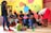 A group of women wearing casual clothing are engaging with children in a colorful room. The walls are painted with bright circles and letters. Some of the women are seated, serving food or interacting with the children, while one stands using a phone. The children are sitting or kneeling on the floor, holding bowls and cups.