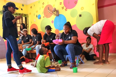A group of women wearing casual clothing are engaging with children in a colorful room. The walls are painted with bright circles and letters. Some of the women are seated, serving food or interacting with the children, while one stands using a phone. The children are sitting or kneeling on the floor, holding bowls and cups.