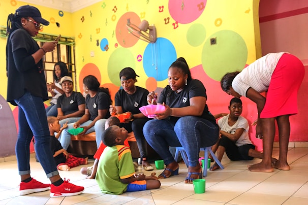 A group of women wearing casual clothing are engaging with children in a colorful room. The walls are painted with bright circles and letters. Some of the women are seated, serving food or interacting with the children, while one stands using a phone. The children are sitting or kneeling on the floor, holding bowls and cups.