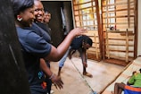 A group of people inside a room engage in cleaning or maintenance activities. One person sweeps the floor with a broom while others stand and observe, smiling or gesturing. The room has wooden structures and a child rests on a bed nearby.