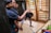 A group of people inside a room engage in cleaning or maintenance activities. One person sweeps the floor with a broom while others stand and observe, smiling or gesturing. The room has wooden structures and a child rests on a bed nearby.