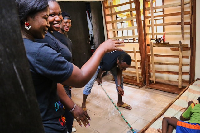 A group of people inside a room engage in cleaning or maintenance activities. One person sweeps the floor with a broom while others stand and observe, smiling or gesturing. The room has wooden structures and a child rests on a bed nearby.