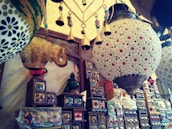 An assortment of colorful decorative items displayed in a shop, including intricately designed ceramic lamps and ornate wooden shelves filled with various decorative boxes. Hanging brass bells add an element of rustic charm.