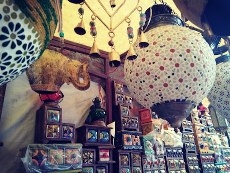 An assortment of colorful decorative items displayed in a shop, including intricately designed ceramic lamps and ornate wooden shelves filled with various decorative boxes. Hanging brass bells add an element of rustic charm.
