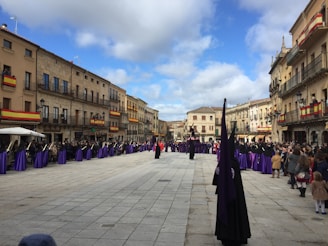 A street procession with participants dressed in purple robes. They are lined up along the sides of a wide stone-paved square surrounded by buildings adorned with Spanish flags. Spectators are gathered to watch the event.