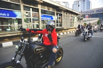 A group of motorcyclists is riding on the road, with the rider in front wearing a red hoodie and a helmet. They seem to be in an urban area with modern buildings and signs visible in the background. The motorcycles are prominent and varied in style, suggesting a casual group ride.
