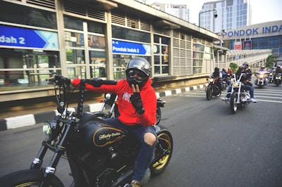 A group of motorcyclists is riding on the road, with the rider in front wearing a red hoodie and a helmet. They seem to be in an urban area with modern buildings and signs visible in the background. The motorcycles are prominent and varied in style, suggesting a casual group ride.