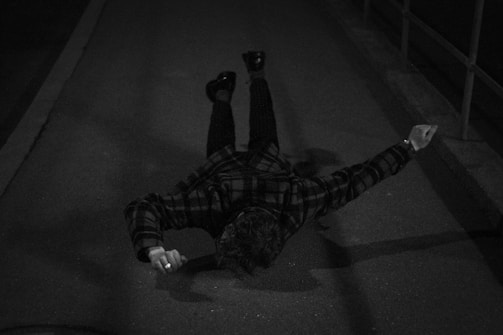 A person lies face down on a pavement in a dark, deserted setting. They are wearing a plaid coat and their arms are stretched out slightly to the sides.