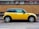 A yellow and black compact car is parked on the side of a street, parallel to a brick wall. The car is a Mini Cooper, characterized by its distinct shape and design. The background features a wall with alternating yellow and black bricks, topped with a row of windows from a neighboring building.