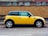 A yellow and black compact car is parked on the side of a street, parallel to a brick wall. The car is a Mini Cooper, characterized by its distinct shape and design. The background features a wall with alternating yellow and black bricks, topped with a row of windows from a neighboring building.