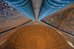 A detailed view of an intricately patterned dome interior featuring geometric designs and calligraphy. The blend of blue and gold tiles adds a rich and artistic atmosphere, with light reflecting off the polished surfaces.