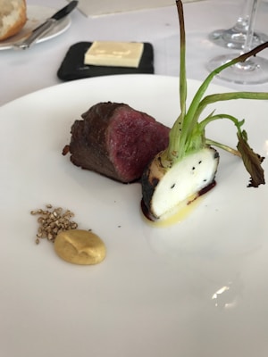 A gourmet dish featuring a piece of medium-rare meat and a root vegetable with charred greens on a white plate. Accompanied by a small dollop of mustard and some seeds. A slab of butter is on a square black dish in the background along with a piece of bread and cutlery.
