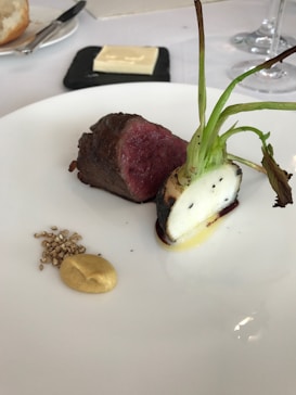 A gourmet dish featuring a piece of medium-rare meat and a root vegetable with charred greens on a white plate. Accompanied by a small dollop of mustard and some seeds. A slab of butter is on a square black dish in the background along with a piece of bread and cutlery.