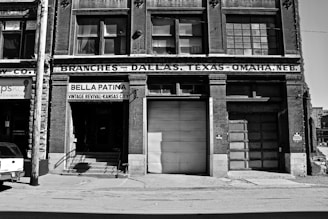 A black and white photograph of an old brick building with signage for 'Bella Patina', a vintage revival store in Kansas City. The structure features large windows and a garage door. The facade displays the text 'Branches – Dallas, Texas – Omaha, Neb.', indicating multiple locations. A power line pole and part of a vehicle are visible in the foreground.