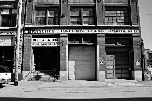 A black and white photograph of an old brick building with signage for 'Bella Patina', a vintage revival store in Kansas City. The structure features large windows and a garage door. The facade displays the text 'Branches – Dallas, Texas – Omaha, Neb.', indicating multiple locations. A power line pole and part of a vehicle are visible in the foreground.