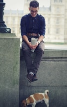A man with glasses, wearing a dark shirt and suspenders, sits on a stone ledge holding a rolled-up newspaper, smiling down at a small dog standing on the ground below. The background features a blurred view of a large building, suggesting an urban setting.