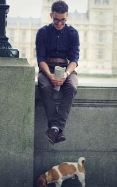 A man with glasses, wearing a dark shirt and suspenders, sits on a stone ledge holding a rolled-up newspaper, smiling down at a small dog standing on the ground below. The background features a blurred view of a large building, suggesting an urban setting.