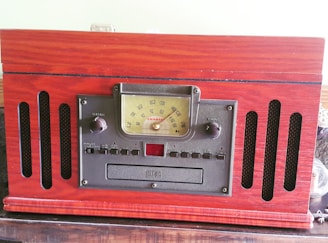A vintage-style radio with a wooden finish featuring a central analog tuning dial and various control knobs on its front panel. The radio has a classic design with speaker grilles on either side.