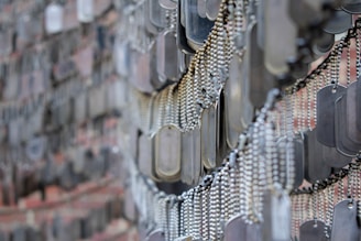 A detailed arrangement of numerous metallic dog tags hanging in close proximity, with each tag attached to a ball chain. The tags create a dense, overlapping pattern, likely part of a memorial or exhibit. The background appears to consist of a blurry brick wall.