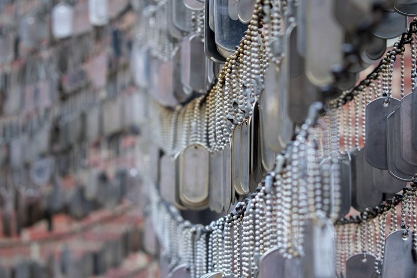 A detailed arrangement of numerous metallic dog tags hanging in close proximity, with each tag attached to a ball chain. The tags create a dense, overlapping pattern, likely part of a memorial or exhibit. The background appears to consist of a blurry brick wall.
