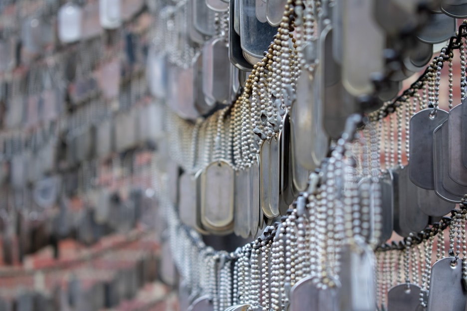 A detailed arrangement of numerous metallic dog tags hanging in close proximity, with each tag attached to a ball chain. The tags create a dense, overlapping pattern, likely part of a memorial or exhibit. The background appears to consist of a blurry brick wall.