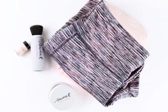 A neatly folded pair of striped athletic shorts is placed alongside a set of beauty products, including a cylindrical container labeled 'Sweat', a makeup brush, and a small jar. The items are arranged on a white surface.