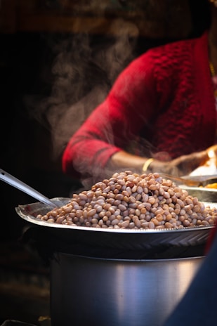 A large tray filled with peanuts is placed on top of a cooking pot, with steam gently rising above it. In the background, a person in a red sweater is partially visible, possibly involved in the preparation or serving process.