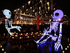 A nighttime display featuring two large illuminated skeleton figures sitting opposite each other, surrounded by a variety of lit candles and decorations. In the background, there are brightly lit buildings adorned with banners, enhancing the festive atmosphere. An orange floral cross stands prominently between the skeletons.