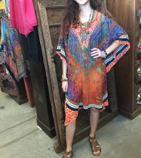 A person wearing a colorful, vibrant kaftan with various patterns stands in a textile shop. The kaftan is predominantly orange, blue, and purple with intricate designs. They are accessorized with colorful beaded jewelry and are in front of a carved wooden panel. Various colorful fabrics hang in the background.