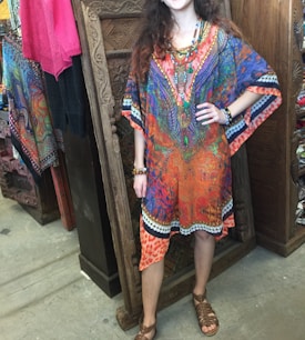 A person wearing a colorful, vibrant kaftan with various patterns stands in a textile shop. The kaftan is predominantly orange, blue, and purple with intricate designs. They are accessorized with colorful beaded jewelry and are in front of a carved wooden panel. Various colorful fabrics hang in the background.