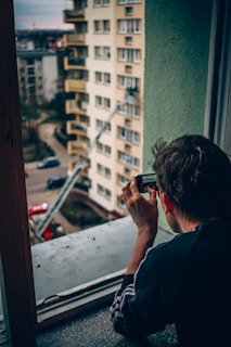 A person is taking a photo with a smartphone from a window ledge, overlooking a scene outside where a fire truck is parked near a tall beige apartment building. A firefighter can be seen on a ladder reaching one of the upper floors of the building.
